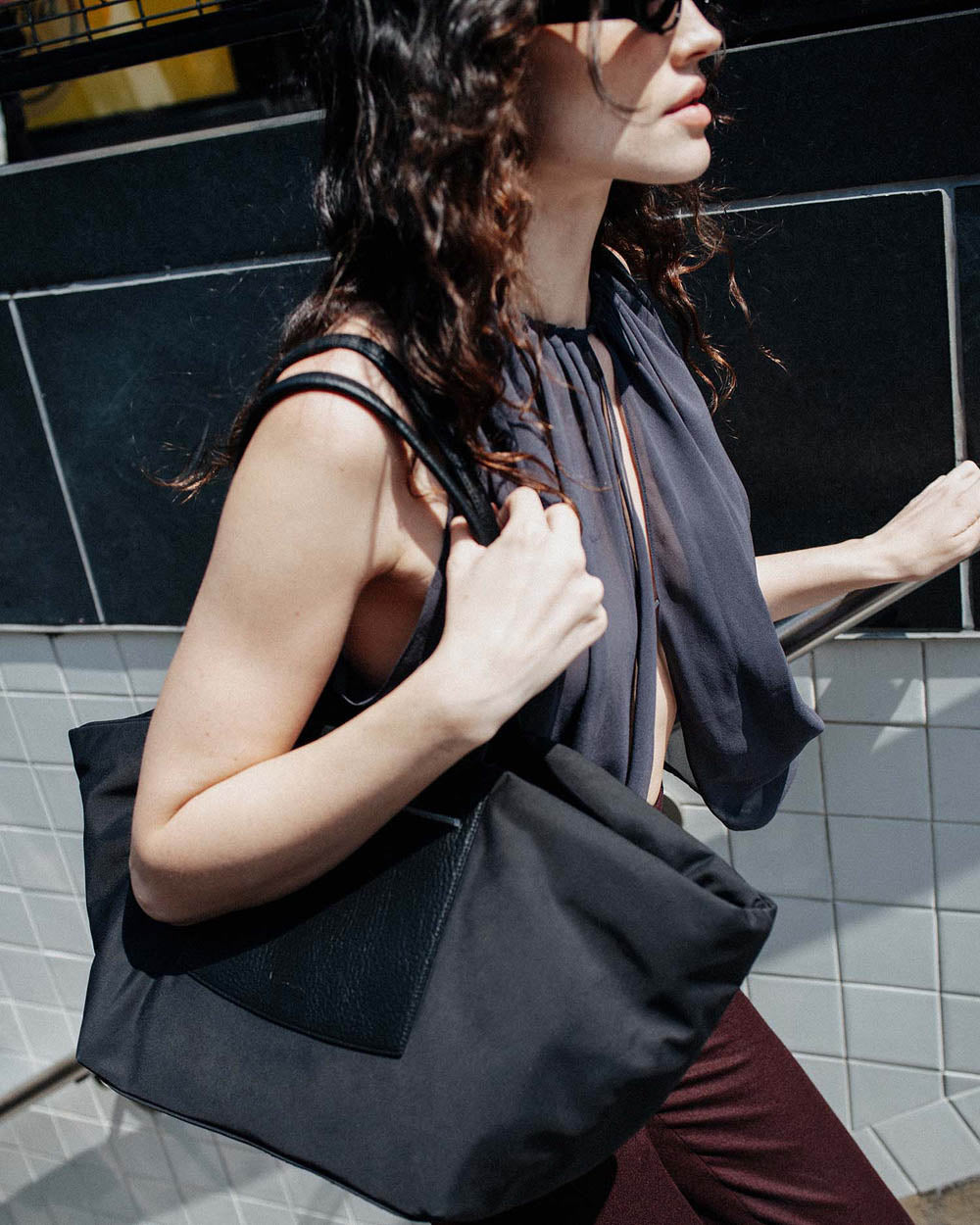 A photo featuring a model wearing black sunglasses and a sheer black sleeveless top stands against a tiled wall, carrying a large black Duo Tote Bag with a leather front pocket.