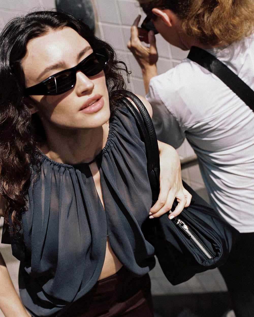 A photo featuring a model wearing black sunglasses and a sheer black sleeveless top stands against a tiled wall, carrying a large black Duo Tote Bag with a leather front pocket.