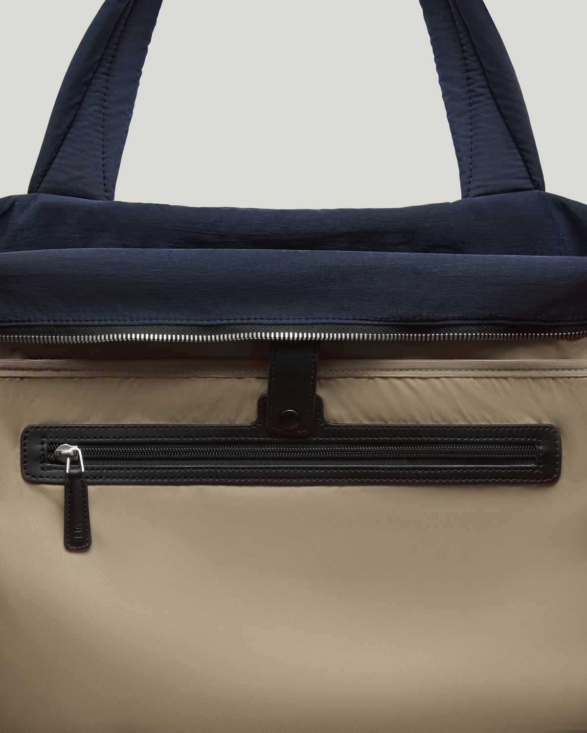 A close-up photo of an Essence Trench Bag featuring a navy blue top handle and flap, a beige body, and a black zippered front pocket with a silver-tone zipper pull.