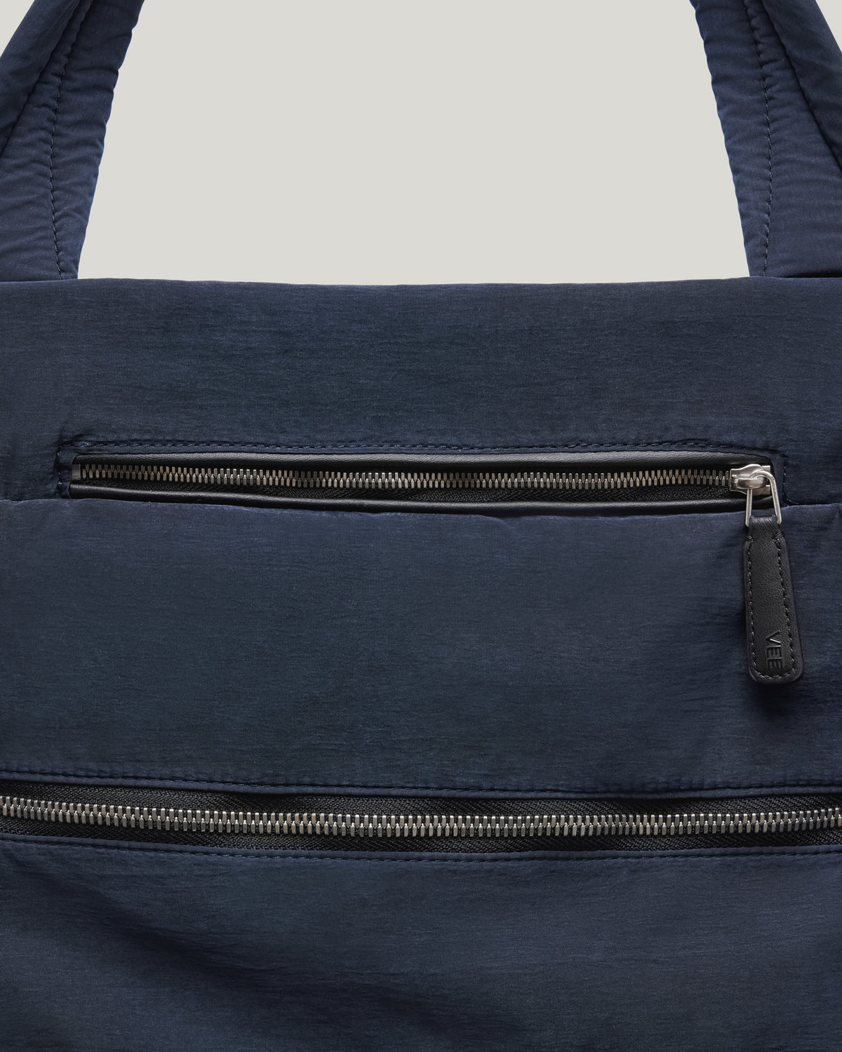 A close-up product photo of an Essence Tote bag featuring two horizontal zippered pockets with dark gray zipper pulls, displayed against a clean white background.