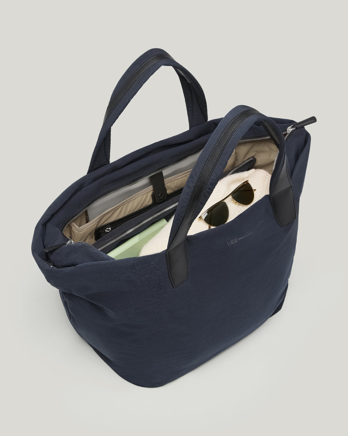 A photo of a navy blue Essence-Trench bag shown open, revealing a beige interior lining with sunglasses, a tablet, and a notebook, set against a solid white background.