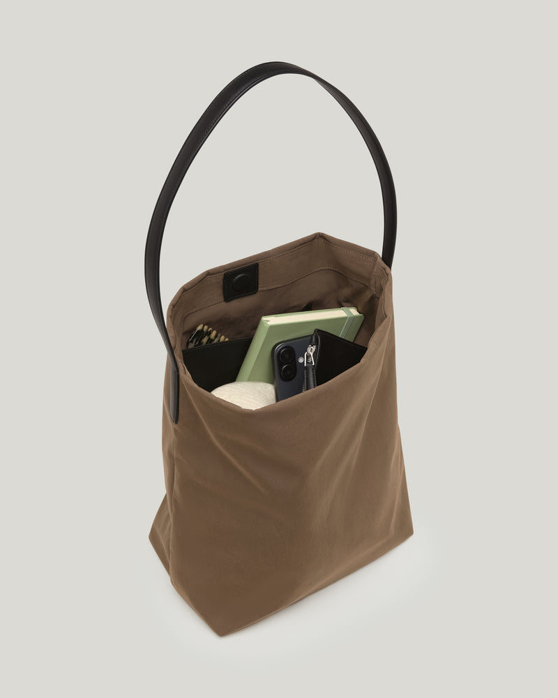 A clean product photo of an Essence vertical tote bag in mink with a black leather handle, shown open and filled with everyday essentials such as a laptop, notebook, and smartphone, displayed against a plain white background.