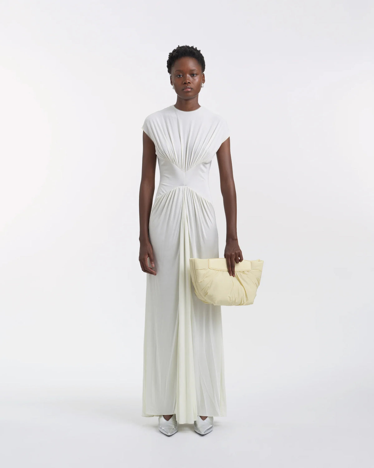 Full-body view of a woman in a white dress holding the Galvan x Vee Collective Caba Clutch Bag in buttermilk with her arm extended in front of her body.