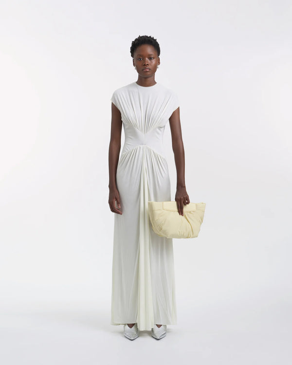 Full-body view of a woman in a white dress holding the Galvan x Vee Collective Caba Clutch Bag in buttermilk with her arm extended in front of her body.
