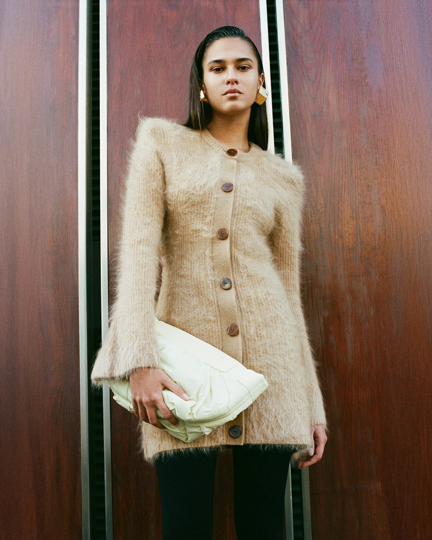 Woman wearing a soft, textured beige cardigan dress, holding the Galvan x VEE Collective Caba Clutch Bag in Buttermilk, styled against a minimalist architectural backdrop.