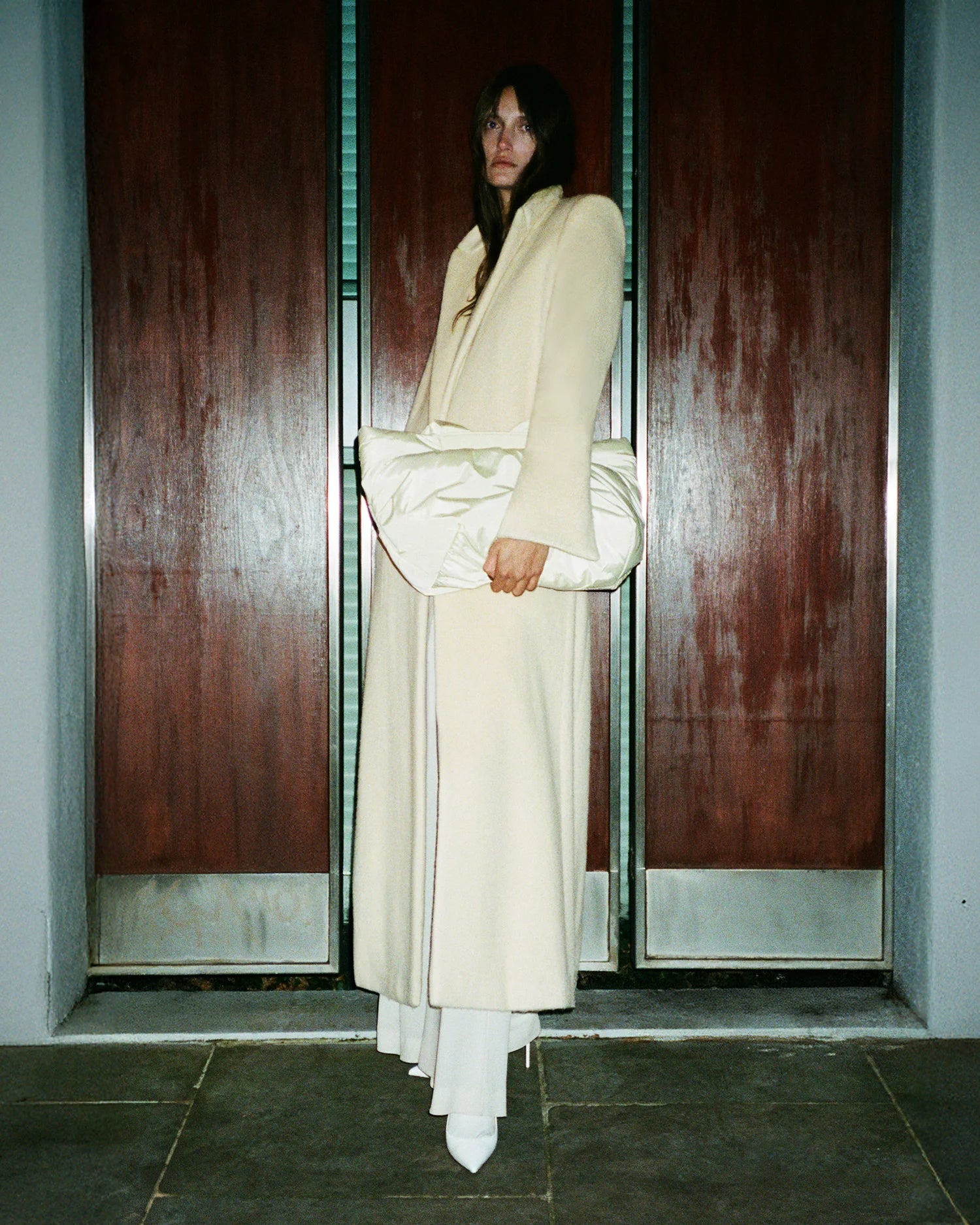 Woman wearing a structured beige coat, holding the GALVAN x VEE Collective Medium Caba Tote Bag in Buttermilk front of a wooden panel backdrop.