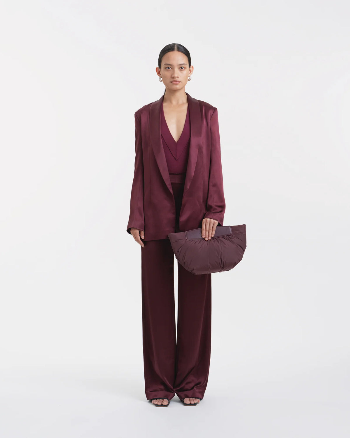 Full-body view of a woman in a mahogany outfit holding the Galvan x Vee Collective Caba Clutch Bag in mahogany with her arm extended in front of her body.