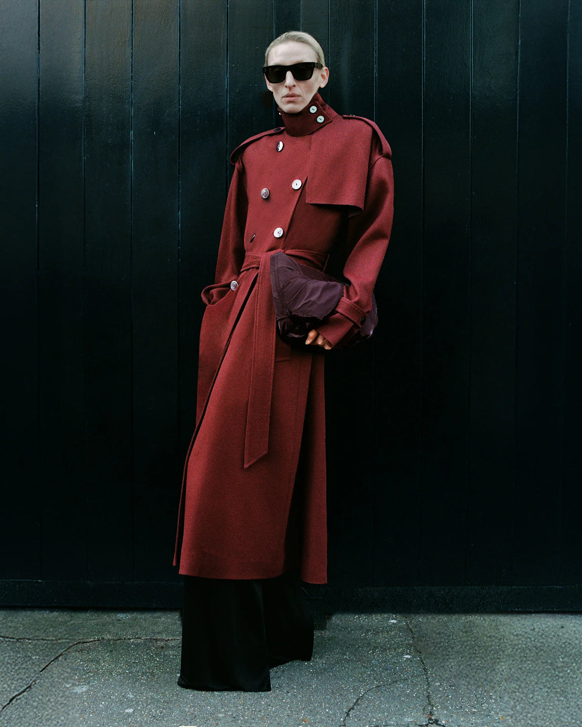 Woman wearing a structured red Coat, holding the GALVAN x VEE Collective Caba Clutch Bag in Mahogany in front of a black wooden panel backdrop.
