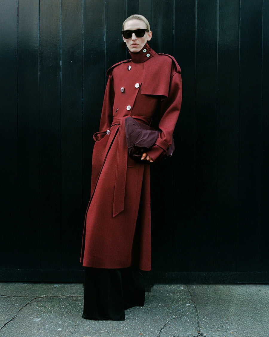 Woman wearing a structured red Coat, holding the GALVAN x VEE Collective Caba Clutch Bag in Mahogany in front of a black wooden panel backdrop.