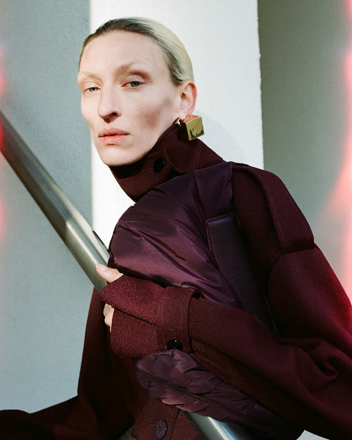 Woman wearing a red coat and geometric gold earrings, holding the GALVAN x VEE Collective Caba Clutch Bag in Mahogany.