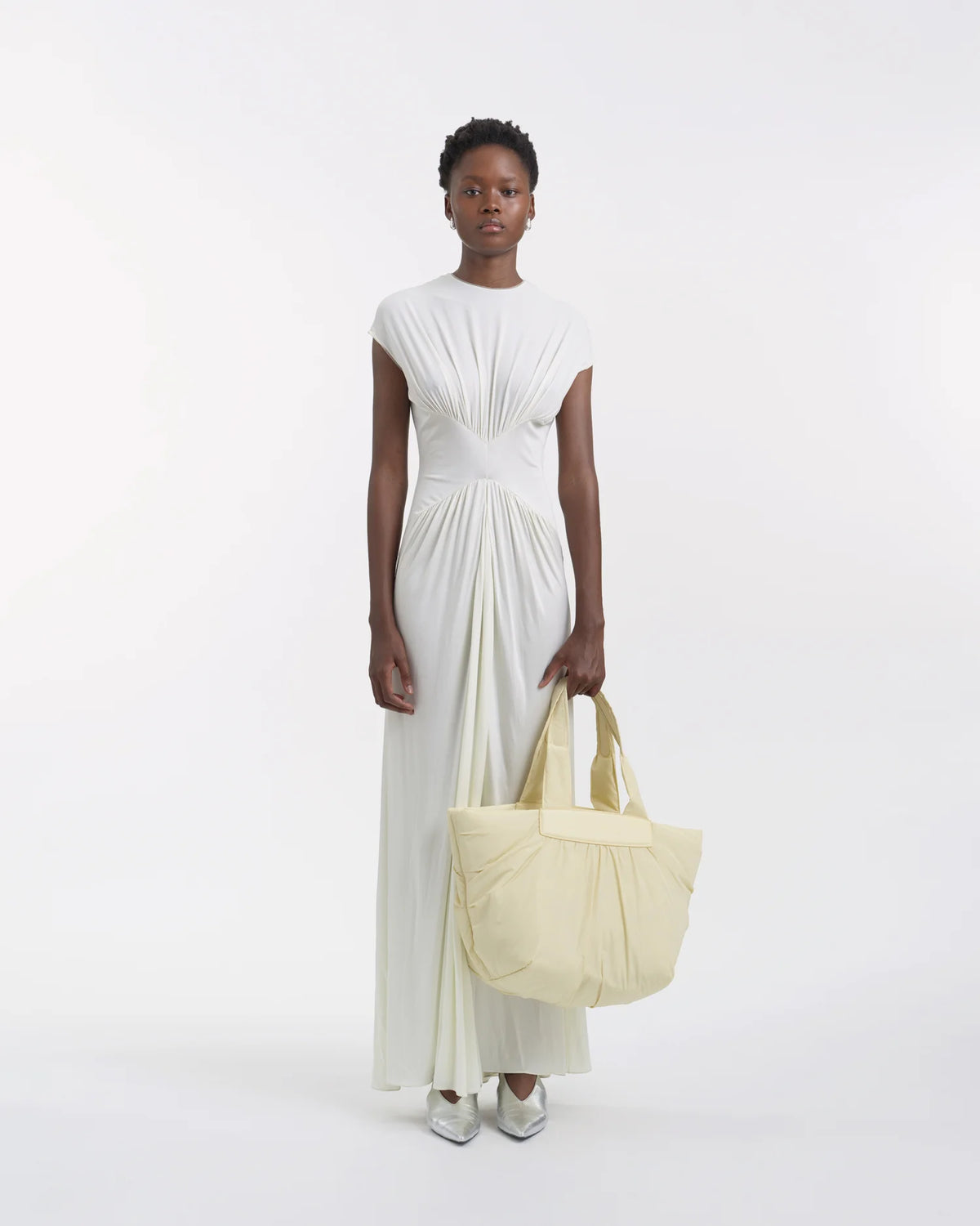 Full-body view of a woman in a white dress holding the Galvan x Vee Collective Caba Medium Tote Bag in buttermilk, with her arm extended in front of her body.