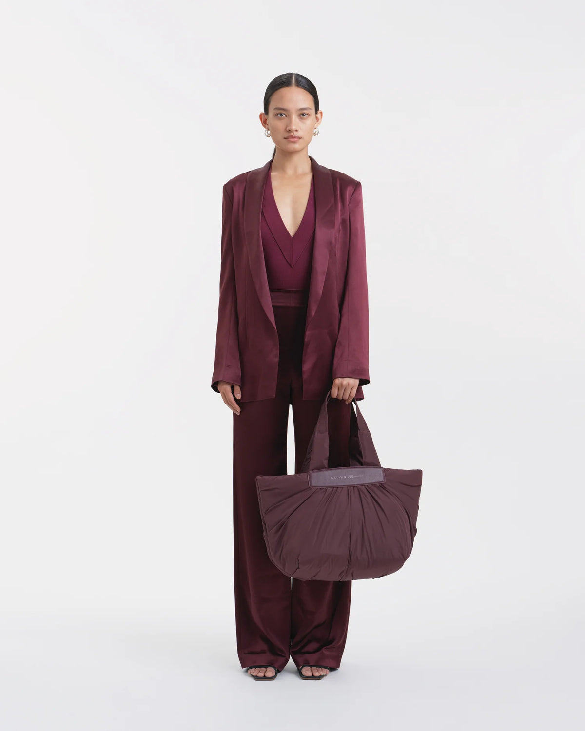 Full-body view of a woman in a mahogany outfit holding the Galvan x Vee Collective Caba Medium Tote Bag in mahogany with her arm extended in front of her body.