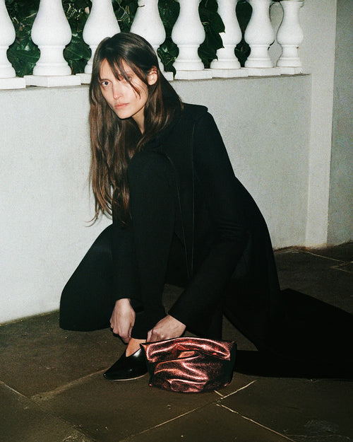 A person wearing an all-black outfit is crouching on a stone floor near a white balustrade, with long hair falling over their shoulders. They are adjusting a black shoe, and the GALVAN X VEE Collective Evening Clutch Bag in Mahogany Metallic rests on the ground beside them