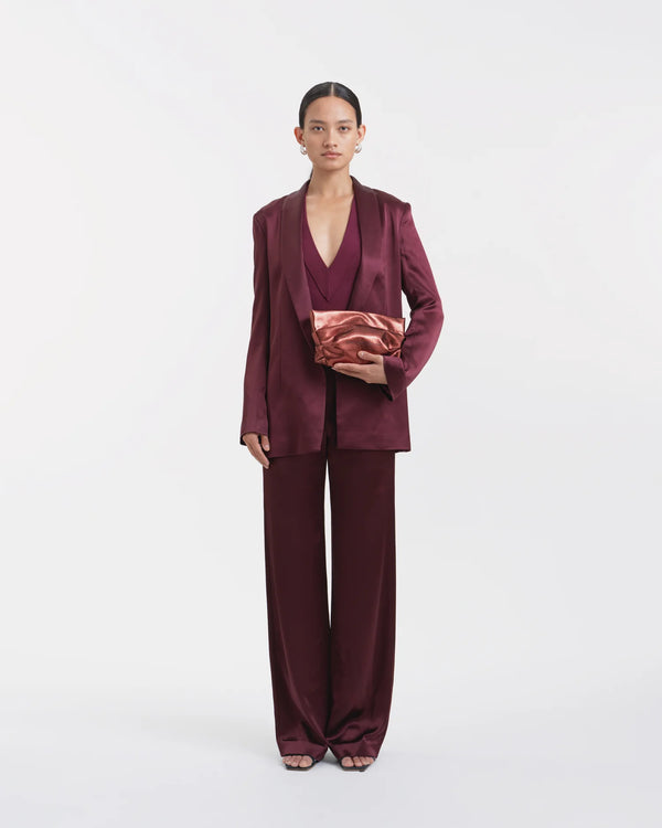 Full-body view of a woman wearing a mahogany outfit, holding the Galvan x Vee Collective Evening Clutch Bag in mahogany metallic in one hand in front of her body.