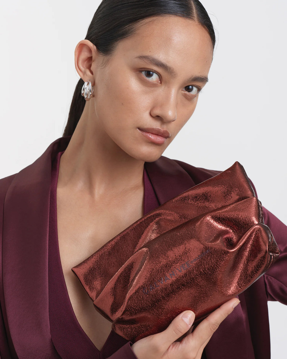 Woman wearing a mahogany outfit, holding the Galvan Evening Clutch Bag in mahogany metallic by Vee Collective in one hand, positioned in front of her upper body at a slight angle.
