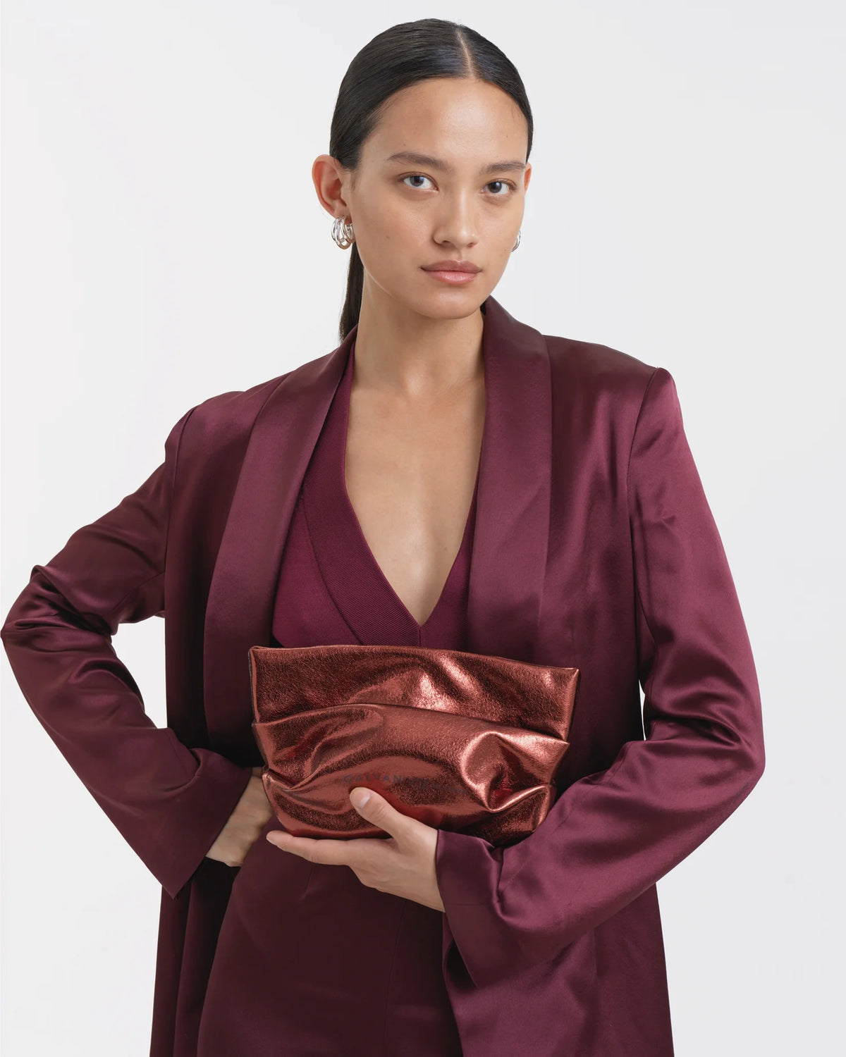 Woman wearing an outfit in mahogany, holding the Galvan x Vee Collective Evening Clutch Bag in mahogany metallic centered in front of her torso.