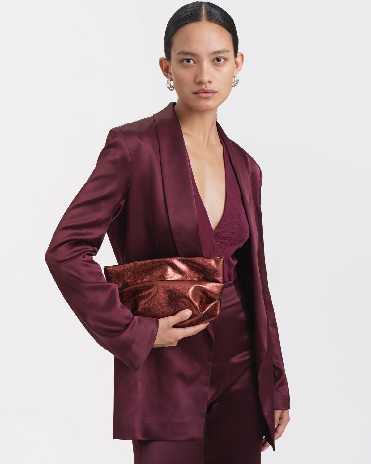 Woman wearing a mahogany outfit holding the Galvan x Vee Collective Evening Clutch Bag in mahogany metallic centered in front of her torso.
