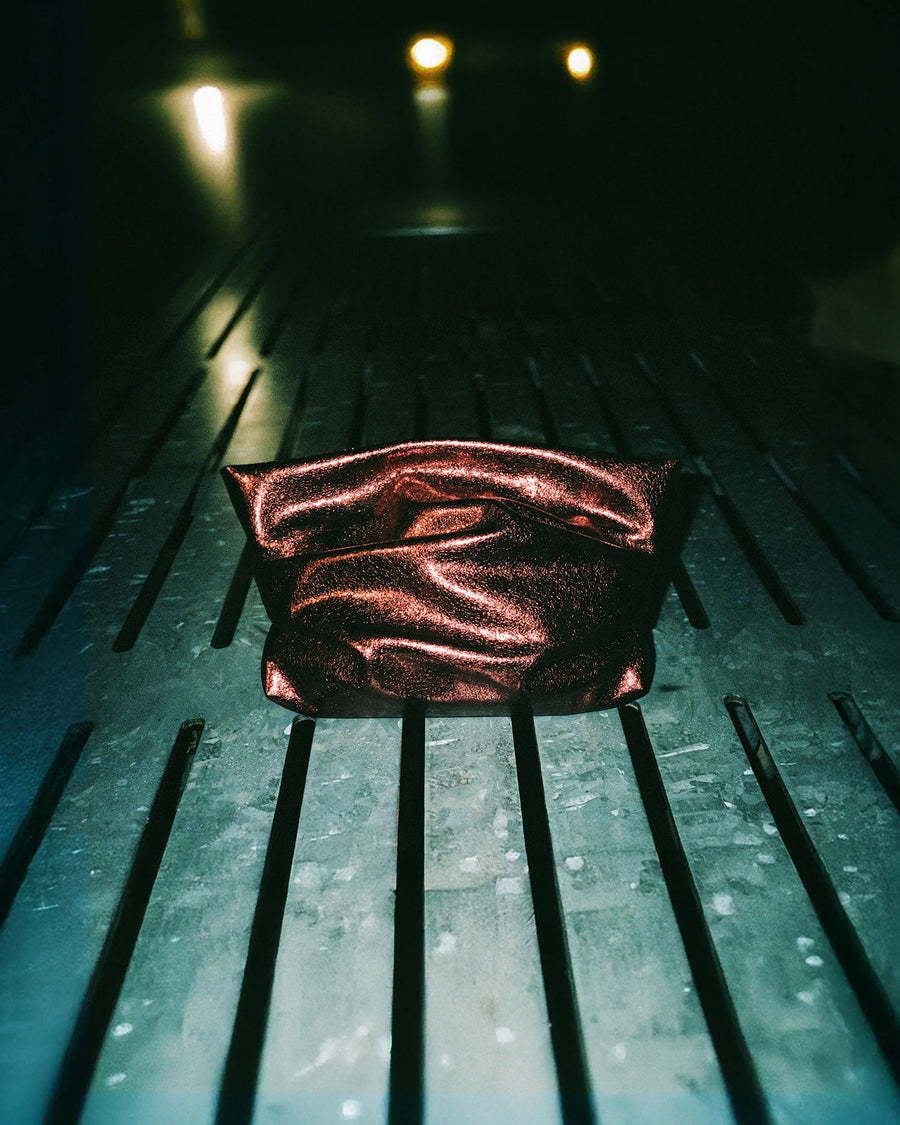 GALVAN X VEE Collectives Evening Clutch Bag in Mahogany Metallic sits centered on a surface with evenly spaced metal slats, illuminated by dramatic overhead lighting in an otherwise dark, industrial-looking environment.