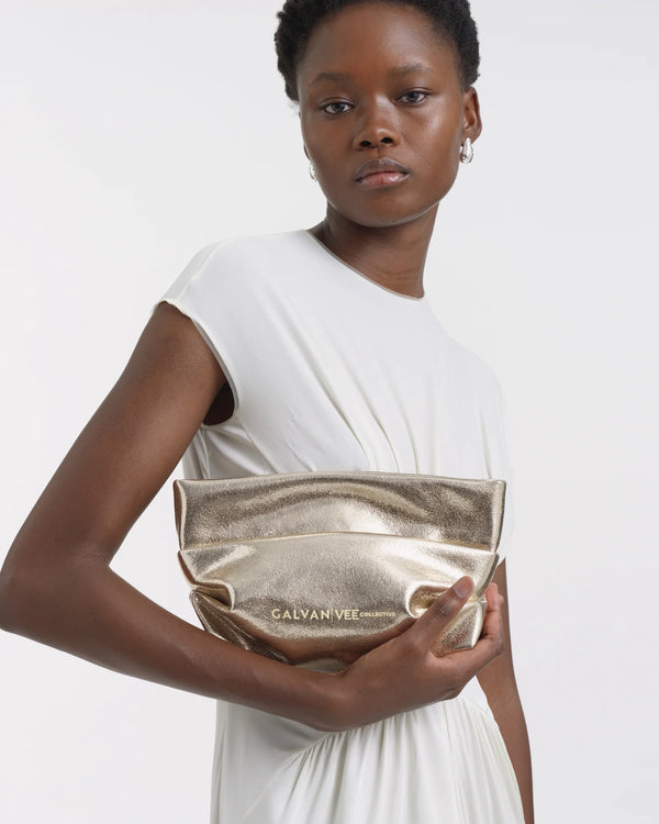 Side view of a woman in a white dress, turned slightly toward the front, holding the Galvan x Vee Collective Evening Clutch Bag in silver metallic centered in front of her upper body.