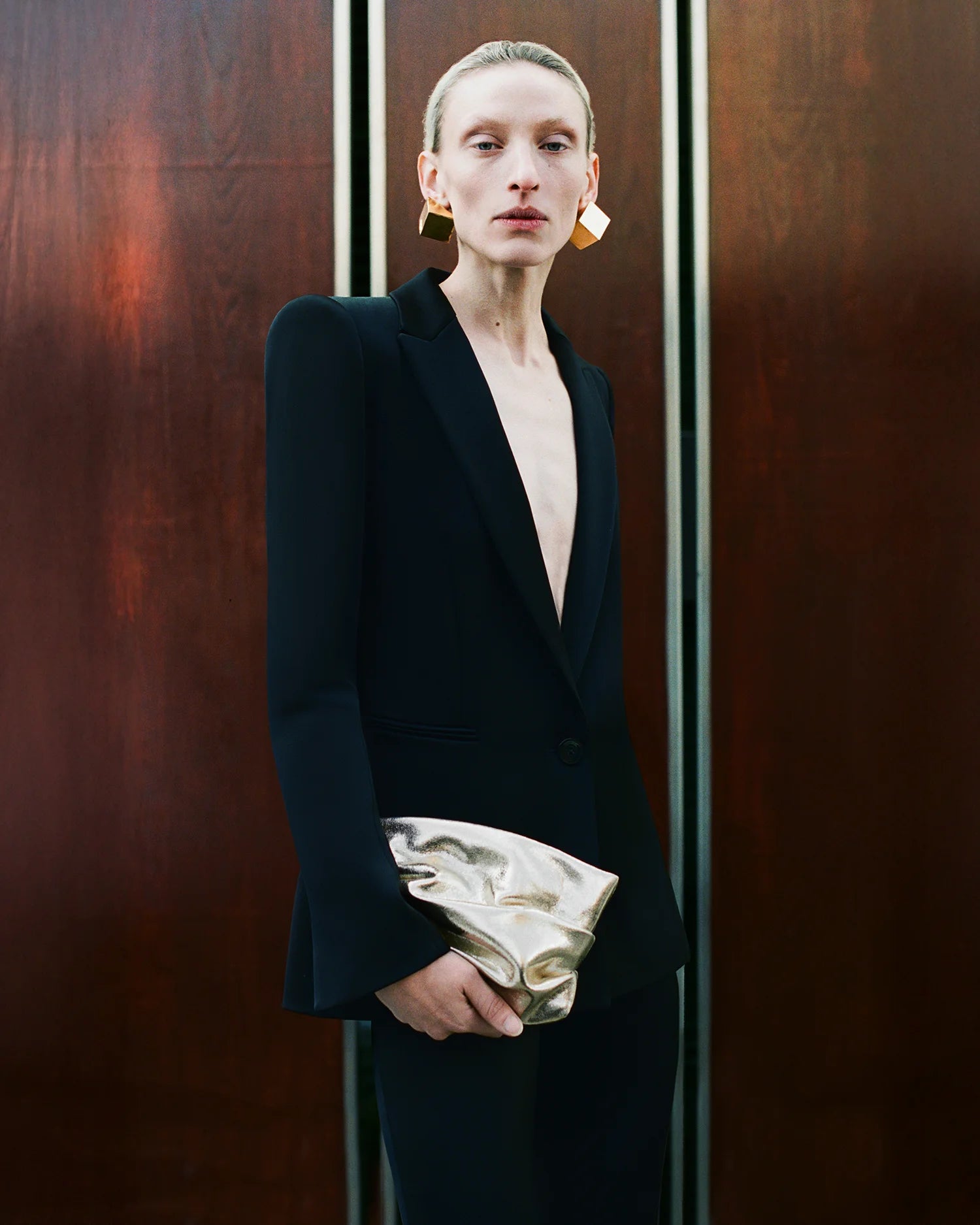 Woman wearing a black blazer and geometric gold earrings, holding the GALVAN x VEE Collective evening Clutch Bag in Gold Metallic in front of a wooden panel backdrop.