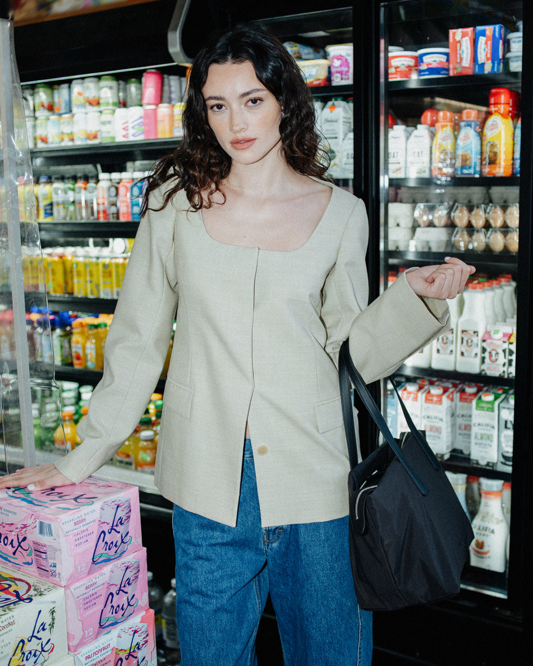 A woman stands in a grocery store aisle wearing a light beige linen-blend jacket with a square neckline and blue jeans, holding a black Essence Tote bag. 