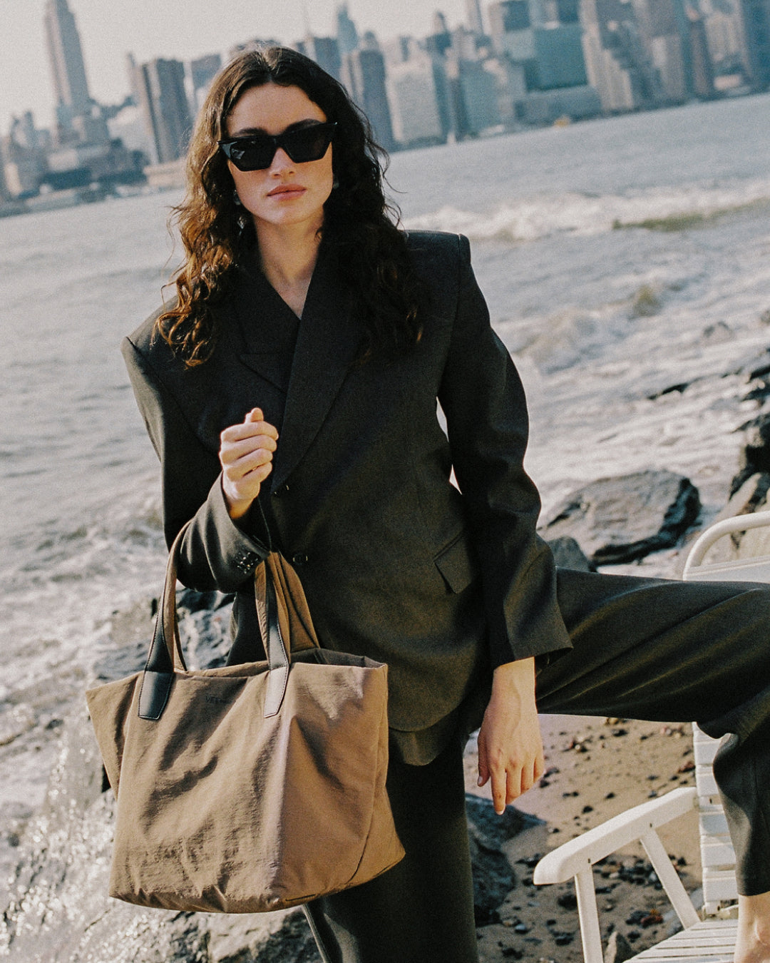 A woman stands on a beach wearing an oversized black blazer and black pants, holding the Essence mink trench bag. The background shows ocean waves and a city skyline.