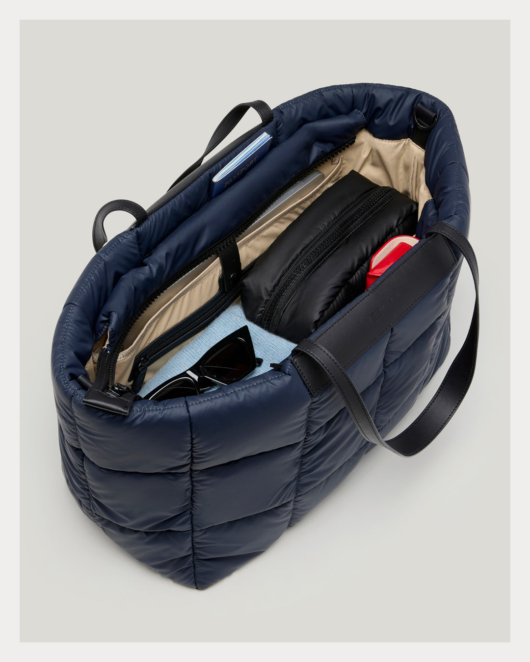 A navy blue puffer tote bag shown open from an overhead angle, revealing a beige interior lining and packed with various items including a black pouch, sunglasses, and a light blue item, set against a solid light gray background.