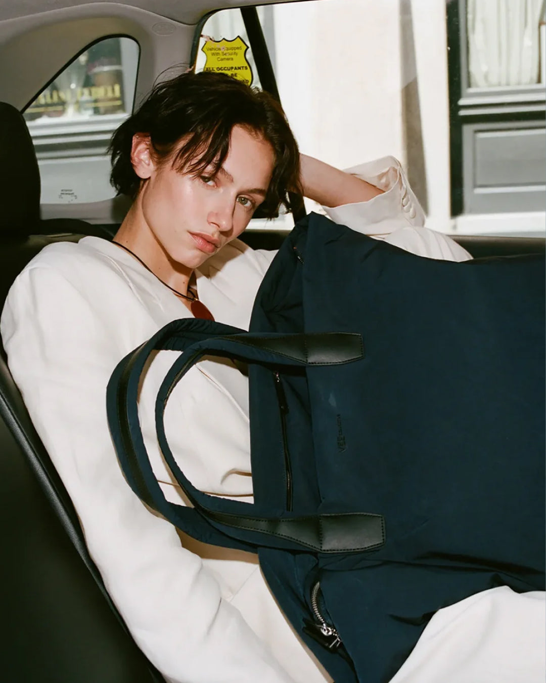 A model sitting in a car backseat wearing a white sweatshirt and holding a large navy blue tote bag with black leather handles.