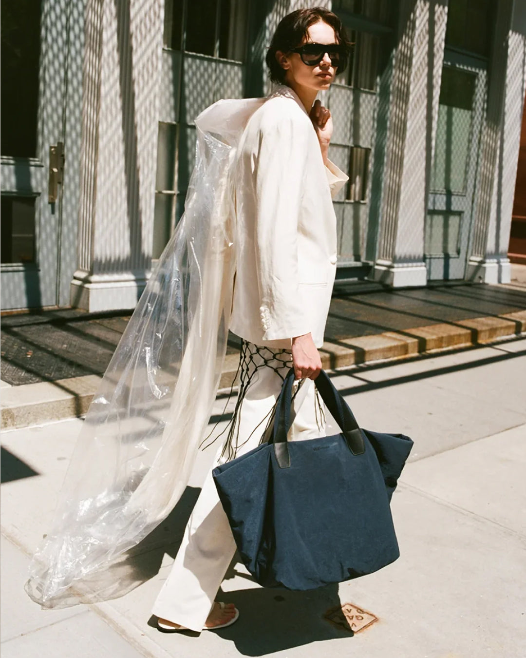 A woman walking outdoors on a city sidewalk carrying a  Essence bag. She is wearing a cream-colored hooded jacket with a long sheer white drape, wide-leg cream pants, and black sunglasses.