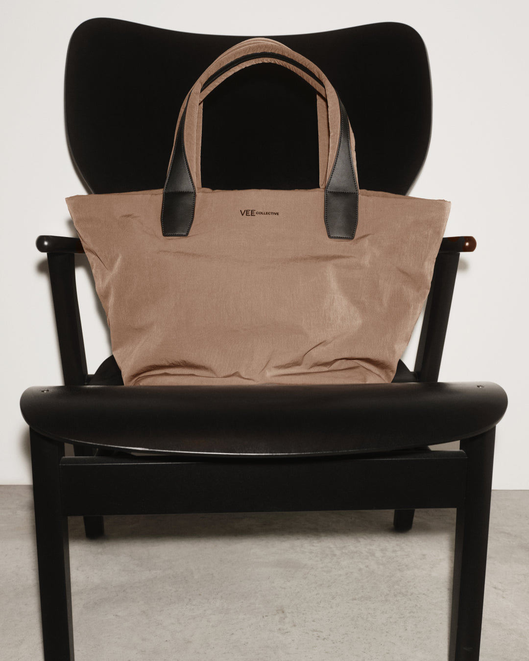 The Essnce City Tote Mink Trench with black leather handles sits on a black wooden chair against a white wall.
