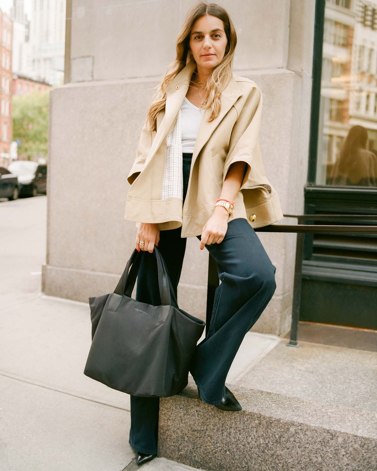 Woman in a beige blazer and dark pants holding a black Essence Tote bag on a city street.