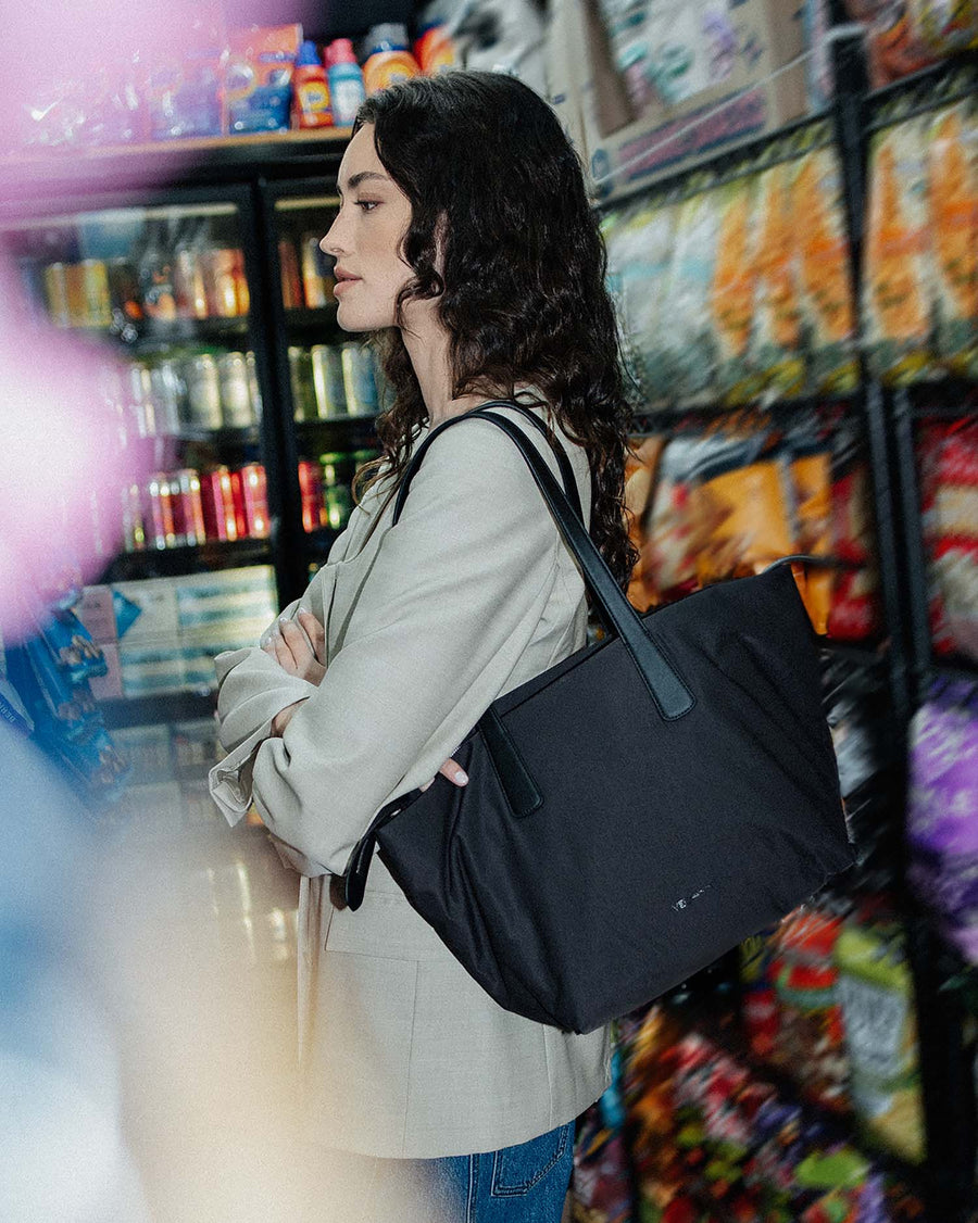 A photo featuring a model wearing a light beige blazer and blue jeans, carrying an Essence City Tote bag with dark teal straps in front of a refrigerator stocked with beverages and dairy products.