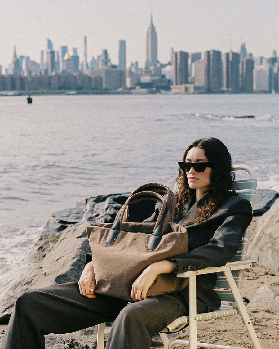 A photo featuring a woman wearing black sunglasses sitting in a beach chair by the water, holding a Mink Essence Tote bag with dark leather handles.