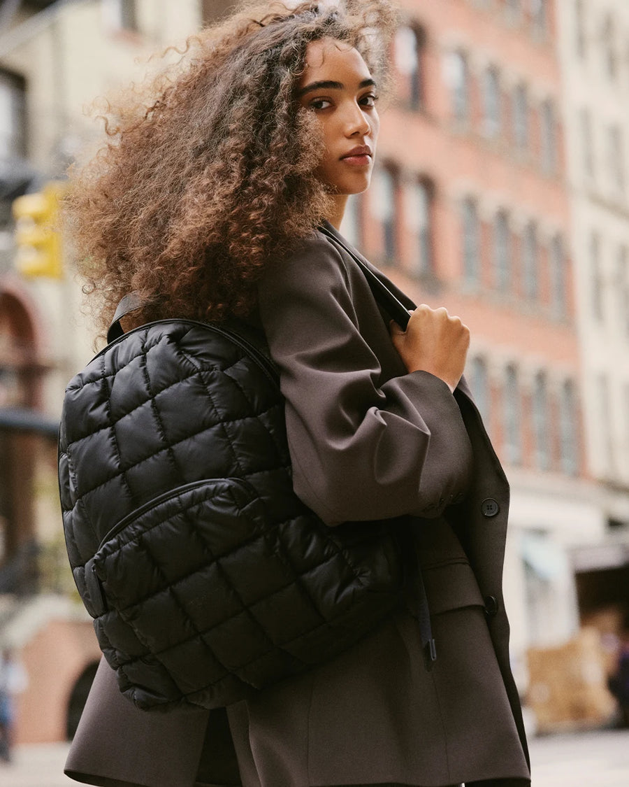 Woman wearing an oversized dark blazer, carrying a quilted Porter Backpack in matt black by Vee Collective over one shoulder, looking back toward the camera on a city street