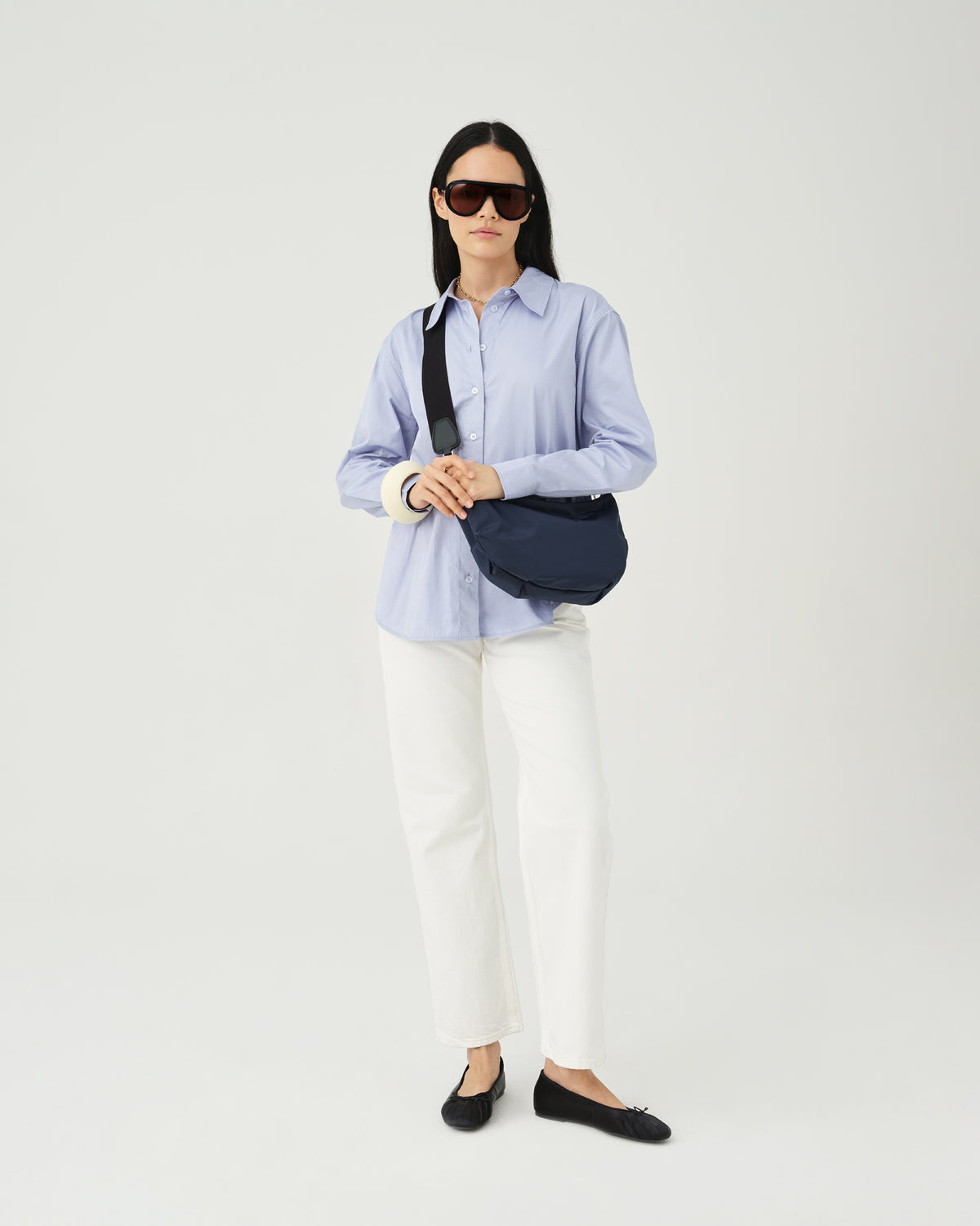 A photo of a woman wearing a light blue button-up shirt, white pants, and dark sunglasses, carrying a Caba Banana Crossbody Bag with a black strap against a plain white background.
