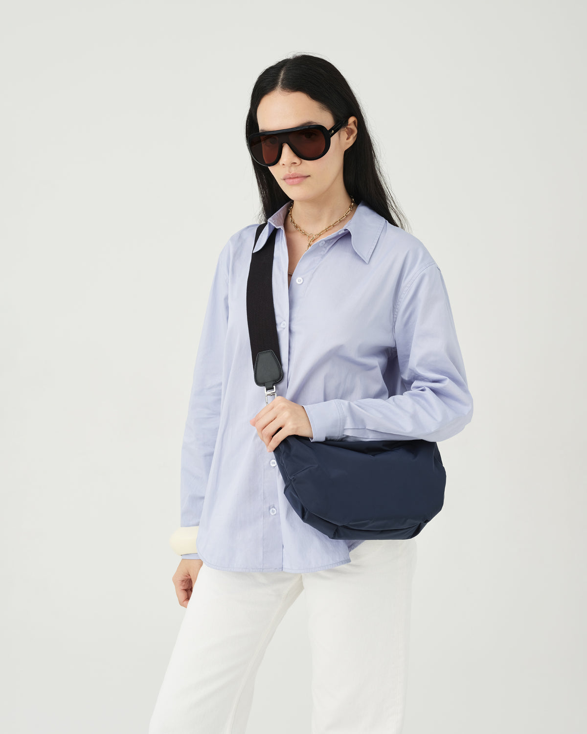 A photo of a woman wearing a light blue button-up shirt, white pants, and dark sunglasses, carrying a Caba Banana Crossbody Bag with a black strap against a plain white background.