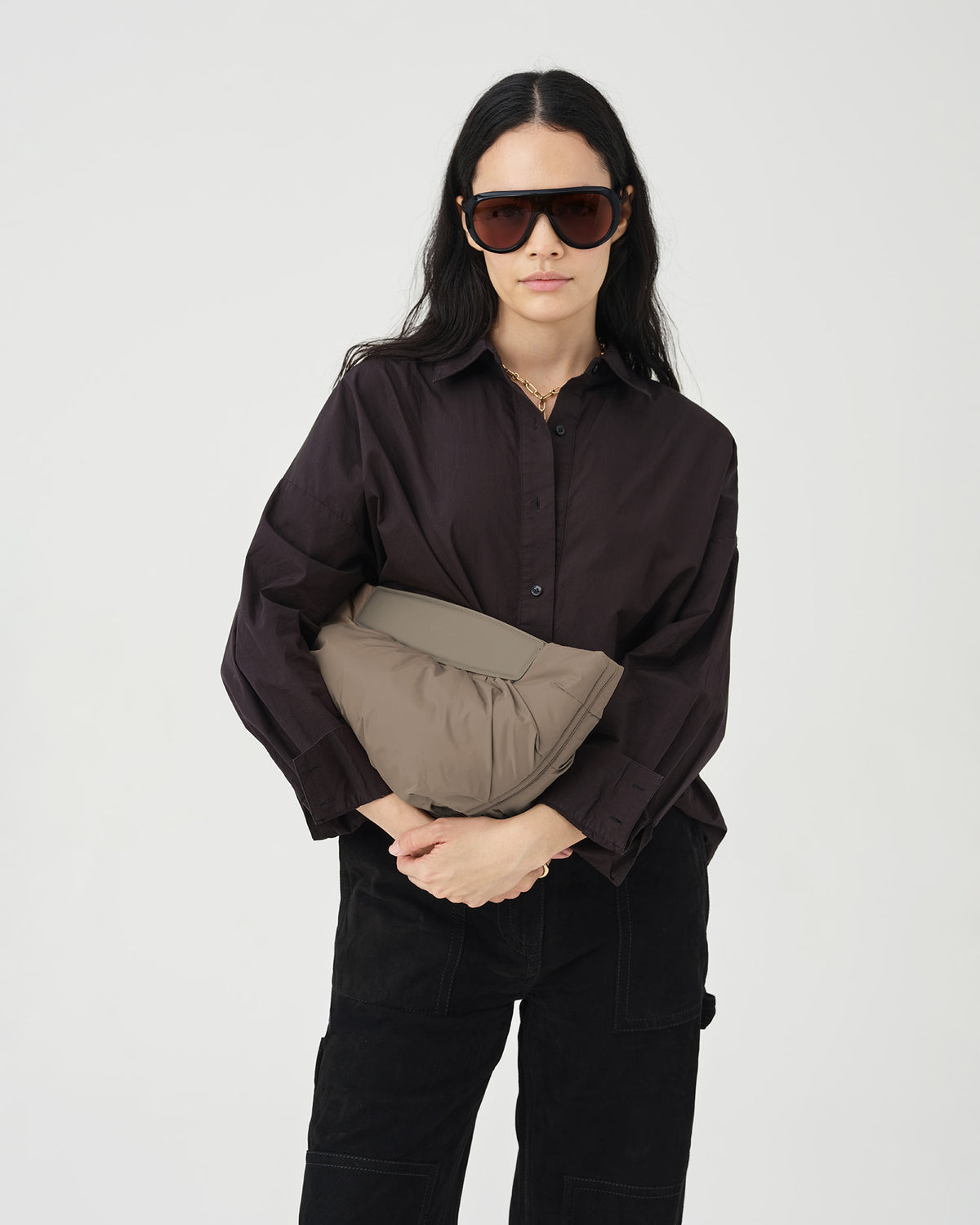 A photo of a model wearing a dark brown button-down shirt, black pants, and oversized dark sunglasses, holding a Caba Clutch Bag against a plain white studio background.