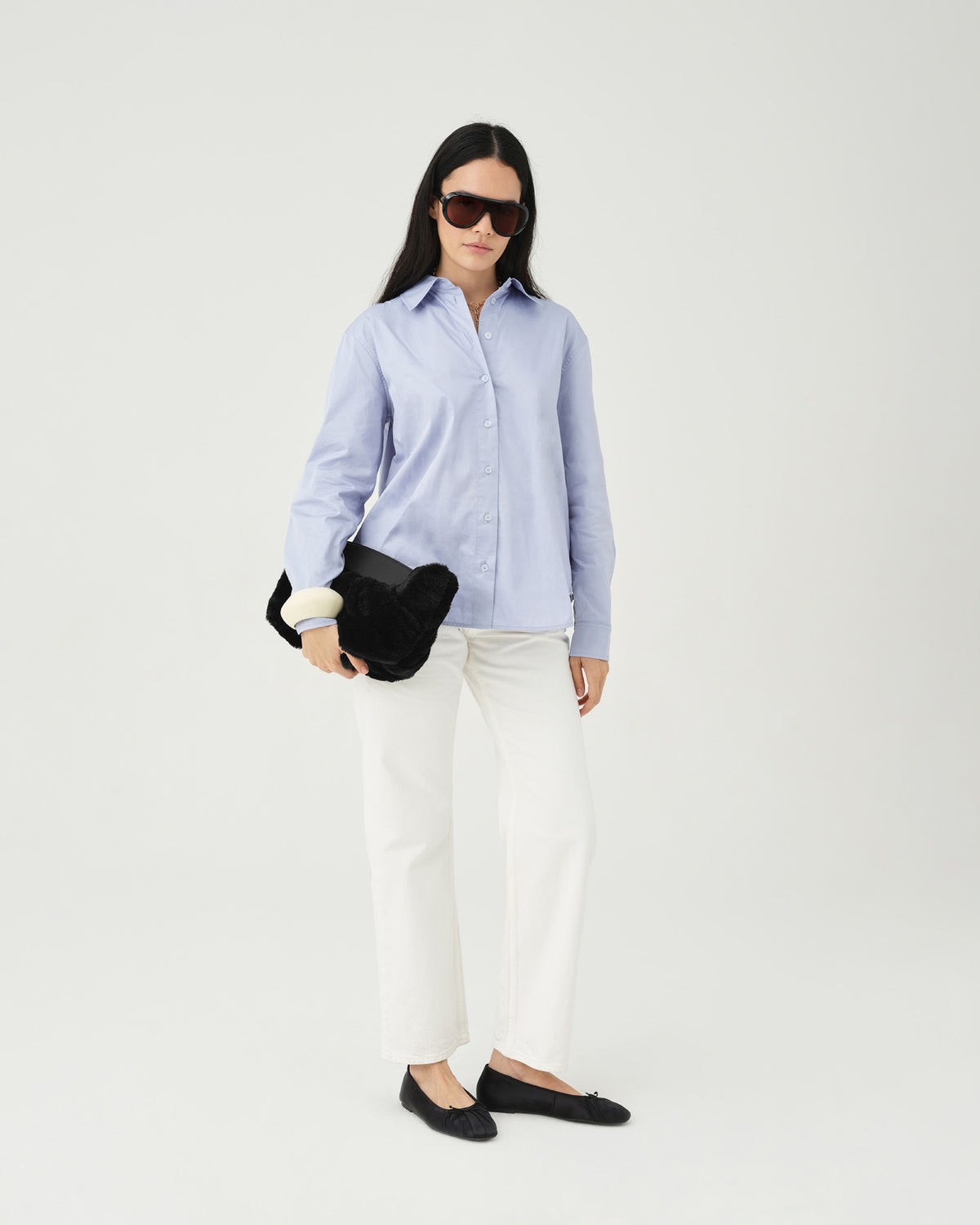 A model wearing a light blue button-up shirt, white pants, and dark sunglasses holds a Caba Fur Clutch with black handles against a white studio background.