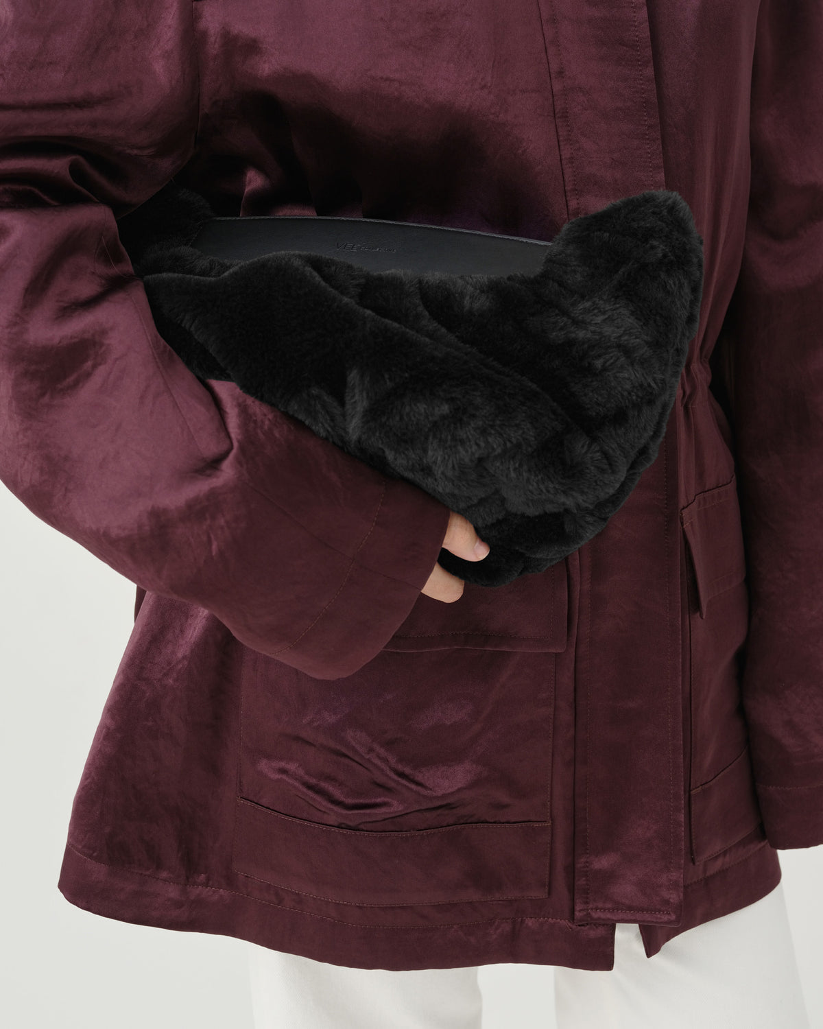 A close-up fashion photo of a model wearing a dark burgundy jacket and holding a black Caba Fur Clutch bag in their hand.