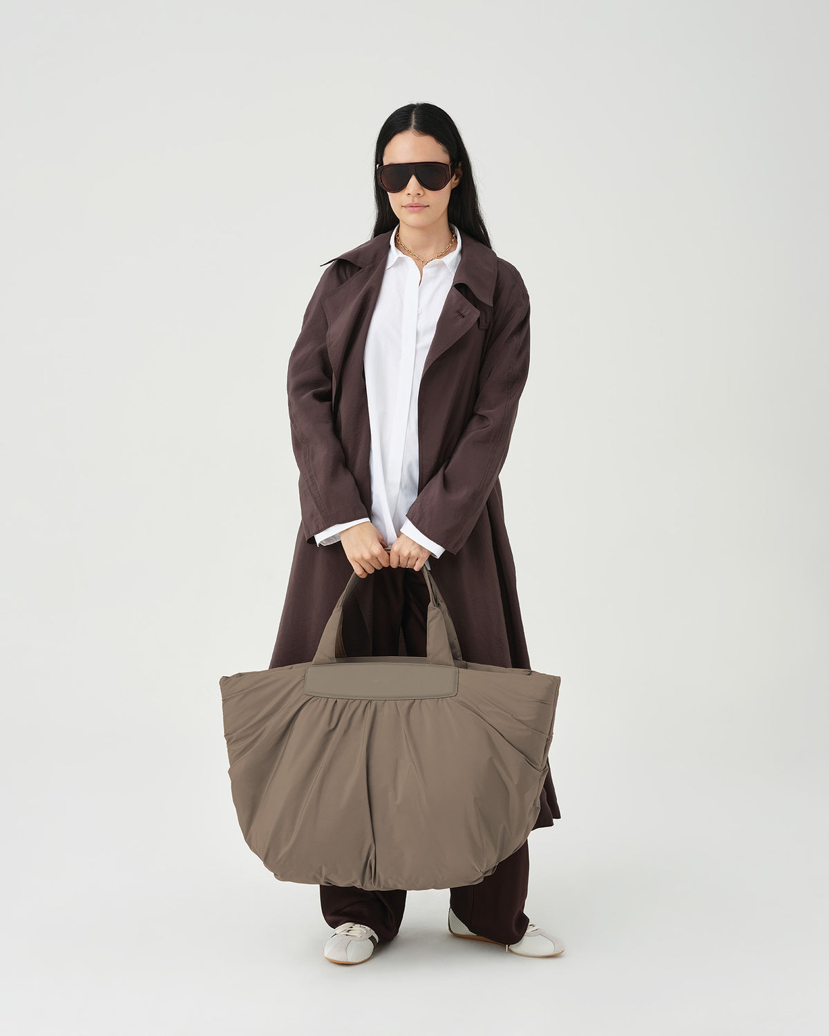 A photo of a model wearing a dark brown oversized coat over a white button-down shirt, dark brown trousers, and dark sunglasses, carrying a Caba Weekender Shoulder Bag against a plain white studio background.