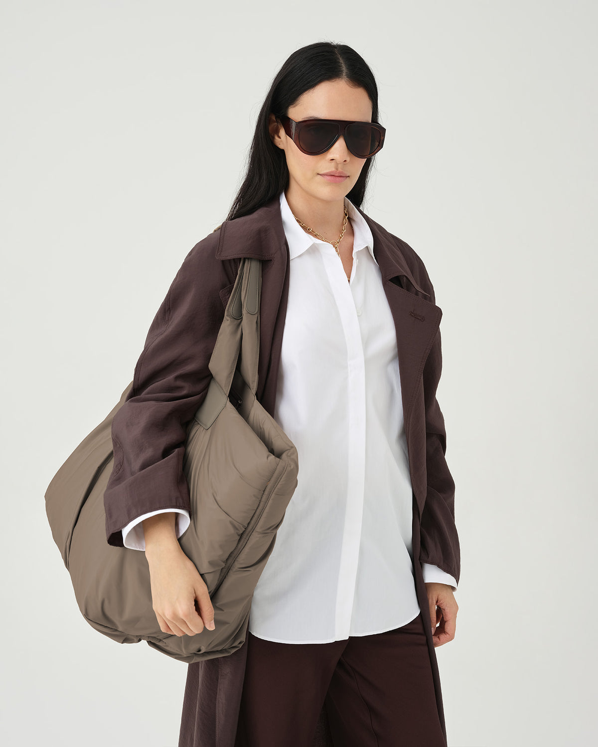 A photo of a model wearing a dark brown oversized coat over a white button-down shirt, dark brown trousers, and dark sunglasses, carrying a Caba Weekender Shoulder Bag against a plain white studio background.