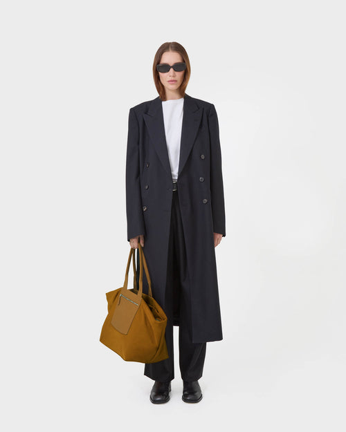A model stands against a white background wearing a long black double-breasted overcoat over a white top and black trousers, holding a duo tote large amber.