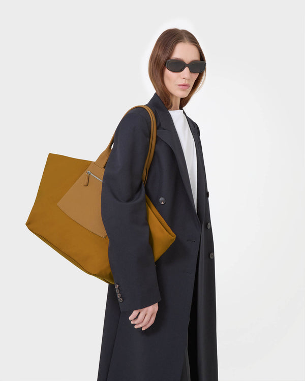 A model wearing a long black coat and sunglasses carries a  duo tote large amber bag with a matching leather accent panel and silver zipper on a light white background.