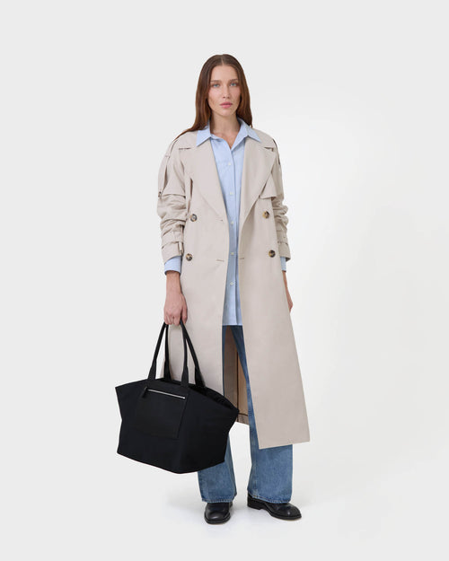 A model stands against a  white studio background wearing a long beige trench coat over a light blue button-up shirt, blue jeans, and black shoes, while holding a large black tote bag.