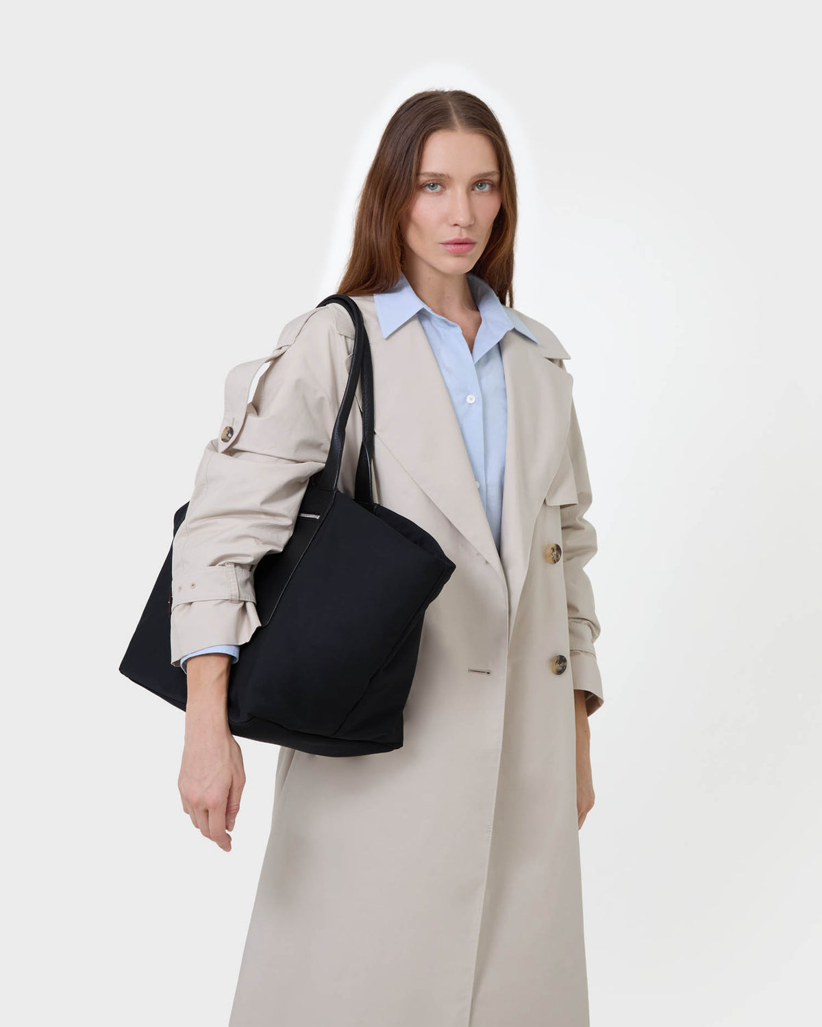A woman wearing a beige trench coat over a light blue collared shirt, carrying a large black tote bag over her shoulder against a plain white studio background.