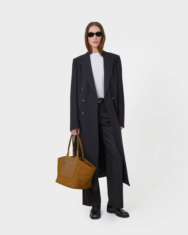 A model stands against a  white studio background wearing a long black  overcoat, black trousers, a white top, black sunglasses, and holding the duo tote medium amber bag.