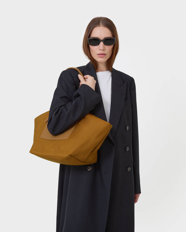 A woman wearing a long black coat, white top, and black sunglasses carries a duo tote medium amber bag with a brown leather accent panel against a white plain  background.