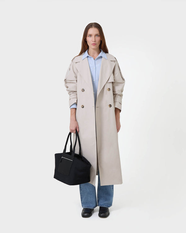 A model stands against a white  studio background wearing a long beige trench coat over a light blue button-up shirt, blue jeans, and black shoes, while holding the black  duo tote bag in medium.