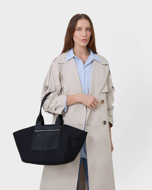 A woman modeling a light beige trench coat over a light blue collared shirt, carrying the  Black Duo Tote in  Medium with a front zip pocket against a white studio background.