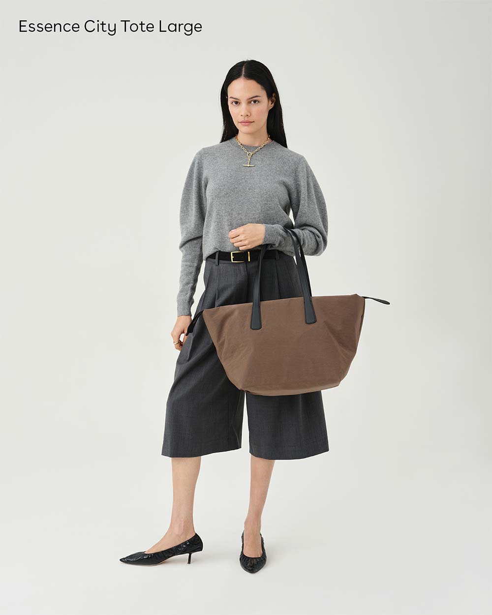 A model stands against a white studio background wearing a gray sweater, dark gray cropped pants, black heels, and holding an Essence City Tote Large bag in mink with black handles. Text in the top left reads 'Essence City Tote Large'.